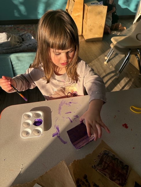 Young child painting a wooden block purple at a table with a small paint palette during an art activity at an early childcare center, with warm sunlight streaming into the classroom.