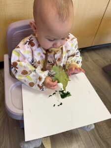 Young girl focused on painting a wooden block purple at a classroom table with a small paint palette, participating in creative art activities as part of early childhood programs in a warm, sunlit learning environment.