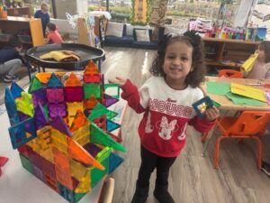 Smiling young girl holding colorful magnetic tiles next to a large structure she built in a bright classroom, showcasing hands-on learning and creativity in the best preschool program.
