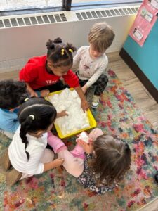 Group of young children sitting on a colorful classroom rug exploring a sensory bin filled with soft material, engaging in hands-on learning at the best daycare for working parents and a nurturing play based preschool environment.