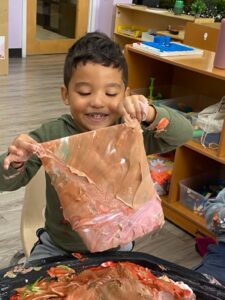 Smiling young child stretching a large piece of textured paint during a hands-on art activity at a day care learning center, highlighting creative exploration in a reggio-inspired preschool classroom environment.